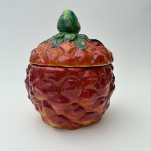 May include: A ceramic lidded jar shaped like a raspberry, featuring a textured, bumpy surface in shades of deep red and orange. The lid is topped with a sculpted green stem and leaves, creating a realistic fruit appearance with a glossy finish.