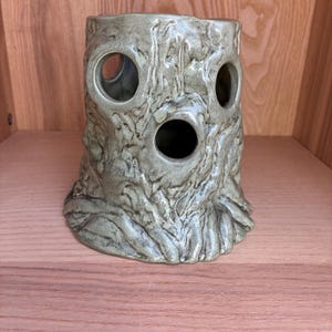 May include: A ceramic vase shaped like a tree stump, with a textured, bark-like surface. The vase has three circular openings and is finished in a muted green-grey glaze. The base has a root-like design.