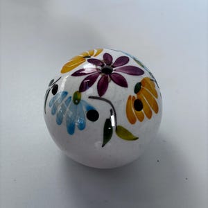 May include: A white ceramic salt shaker with a floral design. The flowers are painted in shades of blue, yellow, orange, and purple. The shaker has three small holes on top for dispensing salt.