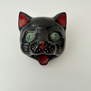 May include: A glossy black ceramic cat head figurine featuring large, pale green eyes, red-painted inner ears, a red nose and mouth area, and small white dots accenting the muzzle. The cat has a wide, open mouth and a smooth, reflective finish.