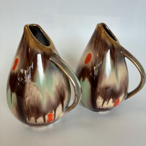 May include: Two teardrop-shaped ceramic jugs with handles. The jugs have a brown, green, and beige glaze with orange and gold accents. The handles and rims are gold. The jugs are likely vintage or retro.