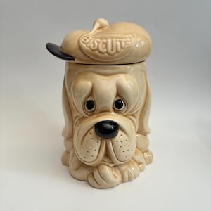 May include: A ceramic biscuit jar shaped like a dog with drooping eyes and a black nose. The dog wears a tan hat with a black brim, featuring the word BISCUITS embossed on the front. The jar has a glossy, light tan finish with darker shading.