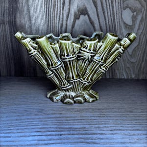 May include: A green ceramic vase styled to resemble bamboo. The vase features a flared, wavy rim and a base designed to look like bamboo stalks. The bamboo detailing includes dark brown accents, giving it a textured look. A decorative item.