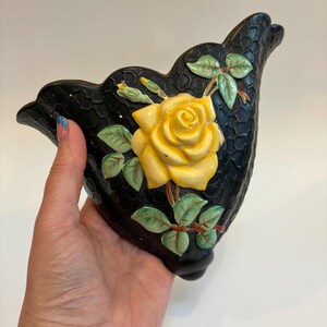May include: Black ceramic wall pocket with a textured surface, featuring a raised yellow rose with green leaves and buds. The decorative floral design adds a touch of vintage charm to any space.