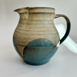 May include: A ceramic jug with a rounded body and a handle. The jug features a speckled, two-tone design with a blue base and a tan upper portion. The handle is dark blue. The jug has a spout for easy pouring.