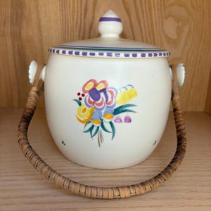 May include: A white ceramic jar with a lid and a woven handle. The jar is decorated with a colourful floral design in yellow, purple, and blue. The lid and rim feature purple and green stripes. The handle is made of woven rattan.