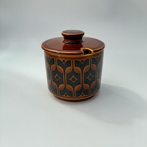 May include: A brown ceramic sugar bowl with a lid. The bowl has a repeating pattern of black and brown shapes. The lid has a small knob and a slot for a spoon. A vintage kitchenware item.