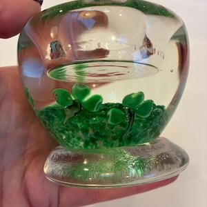 May include: A clear glass votive candle holder with green shamrock accents. The base of the holder is filled with green speckled glass, and the shamrocks are three-leafed clovers. The holder is round with a flat base.