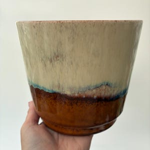 May include: A ceramic planter featuring a two-tone glaze with a creamy off-white upper section that transitions into a deep, glossy brown base. A thin, distinct band of blue-toned glaze separates the two colours, creating a layered effect.