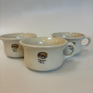 May include: Three off-white ceramic cups with handles. Each cup features a brown and blue eye graphic with the words "WON'T TELL" printed below. The cups have a rounded shape and a small handle.