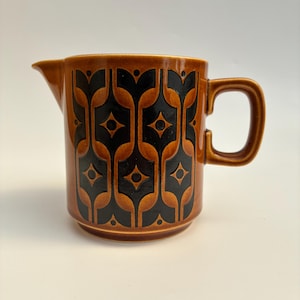 May include: A ceramic creamer jug featuring a warm brown base colour and a repeating black geometric pattern. The jug has a side handle, a small pouring spout, and a cylindrical body with a slightly flared rim and a flat base.