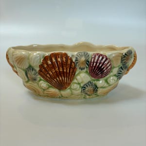 May include: Oval ceramic planter with a cream base, decorated with detailed seashell designs in shades of brown, burgundy, and grey. The exterior features a textured, raised relief pattern of shells and small stones, creating a coastal theme.