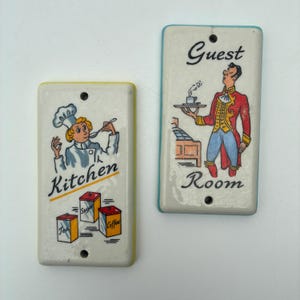 May include: Two rectangular ceramic signs. One sign has a yellow border and features a chef with the word "Kitchen" below. The other sign has a blue border and depicts a waiter with the words "Guest Room". Both signs have a hole at the top for hanging.