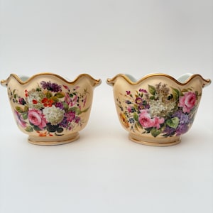 May include: Two cream-coloured porcelain bowls with scalloped, gold-trimmed rims. Each bowl features a colourful floral arrangement including pink roses, white hydrangeas, and purple blossoms painted on the exterior surface.