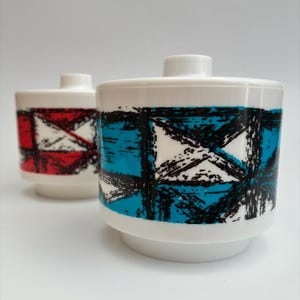May include: Two white ceramic lidded containers with a cylindrical shape and a stepped base. The foreground container features a bright blue and black geometric grid pattern, while the background container displays a red and black version.
