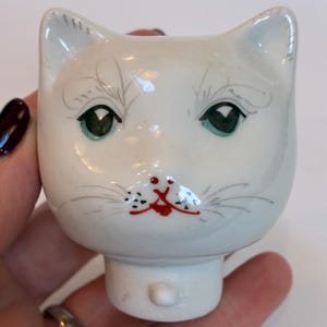 May include: A white ceramic cat head figurine with green eyes, red lips, and grey whiskers. The cat's face is hand-painted with delicate details. The figurine has a small, cylindrical base.