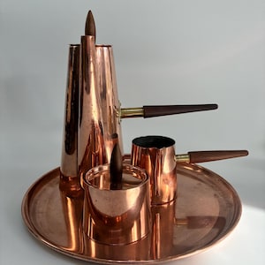 May include: A copper coffee set on a tray. The set includes a tall, conical coffee pot, a small creamer, and a small pot, all with wooden handles. The set is displayed on a round copper tray.