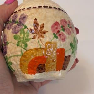 May include: A cream ceramic jar with a lid, adorned with hand-painted floral and geometric patterns. The jar showcases purple, pink, and green flowers, alongside brown and orange geometric shapes. A brown dotted line encircles the lid.