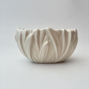 May include: A cream-coloured ceramic bowl featuring a textured exterior with a repeating pattern of overlapping, elongated leaf-like shapes. The bowl has a scalloped, uneven rim that follows the contours of the raised, vertical petal designs.