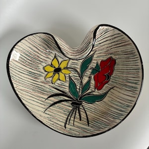 May include: Heart-shaped ceramic dish with a cream background and a black rim. It features a floral design with a yellow flower, a red flower, and green leaves. The background has a striped pattern.