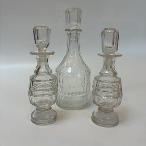 May include: Three clear glass bottles with stoppers. The bottles vary in shape and size, with one tall, slender bottle in the centre and two smaller, decorative bottles on either side. The stoppers are also clear and have a rectangular shape. The bottles are on a white background.