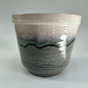 May include: A ceramic plant pot with a gradient design. The top rim is a light beige, transitioning to a dark grey base. A wavy dark blue and black line separates the colours. The pot has a textured, speckled appearance.
