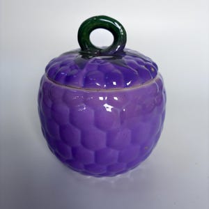 May include: A ceramic jar with a lid, designed to look like a purple grape. The jar has a textured surface and a green loop handle. The jar is a decorative item, suitable for storage.