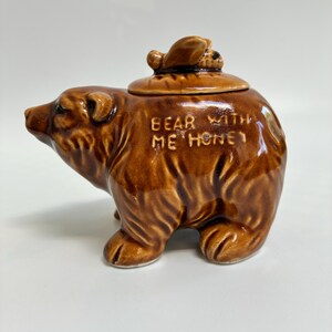 May include: A brown ceramic bear-shaped honey pot with a lid. The words "BEAR WITH ME HONEY" are embossed on the side. The bear is standing on all fours, with a detailed face and fur texture.