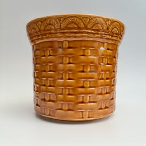 May include: A cylindrical ceramic planter with a glossy, honey-brown glaze. The exterior features a moulded, raised texture that mimics the appearance of a woven basket weave pattern, topped with a decorative border of repeating semi-circular arches.