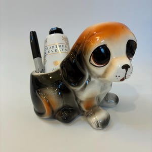 May include: A ceramic puppy-shaped desk tidy in brown, black, and white. The puppy has large eyes and holds a pen and a bottle of lotion. The organizer is designed to hold small items, such as pens and pencils. The lotion bottle is labeled.
