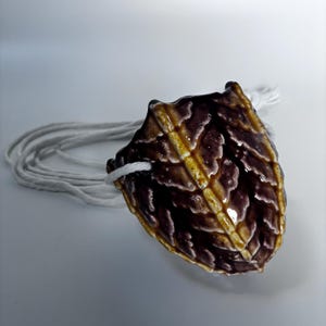 May include: A ceramic cone-shaped ornament with a textured surface in shades of brown, yellow, and white. A white cord is attached. The object is likely a decorative item. The measurements are in centimetres.