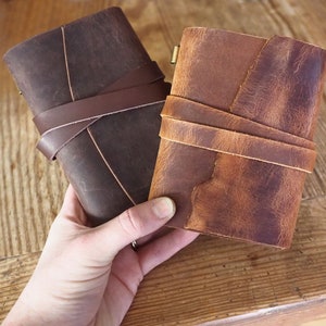 May include: Two leather-bound journals with brown and tan covers. The journals have leather straps that wrap around them and are tied with a knot.