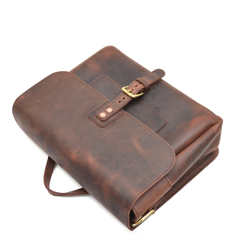 May include: A brown leather satchel with a flap closure and a buckle. The bag has a handle and a shoulder strap. The leather has a distressed look. The hardware is gold-coloured.