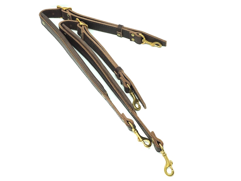 May include: A brown leather camera strap with two brass hooks and a double strap design.