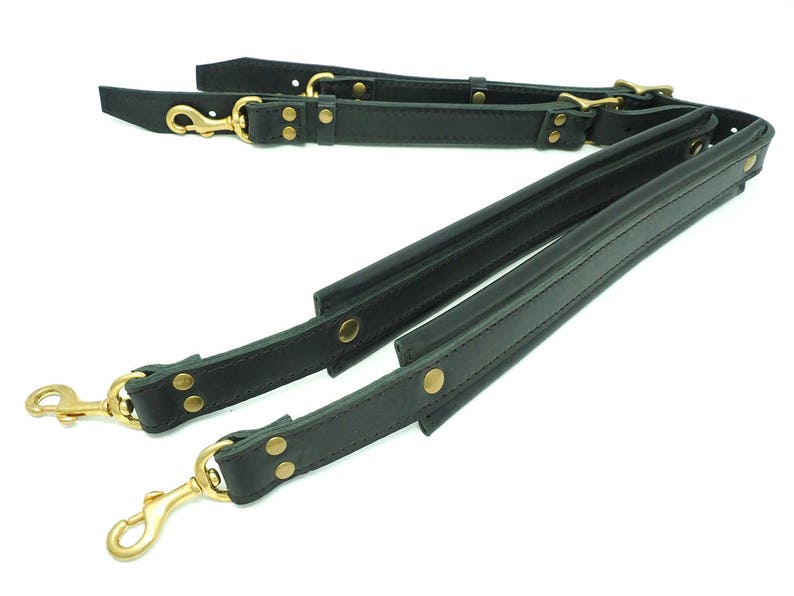 May include: Two black leather straps with brass hardware and clasps. The straps are connected together and are designed to be used as a bag strap.