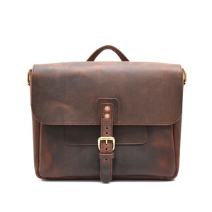 May include: A brown leather satchel with a flap closure and a handle. The bag features a front pocket, a buckle closure, and copper rivets. The leather has a distressed appearance, and the hardware is gold-toned.
