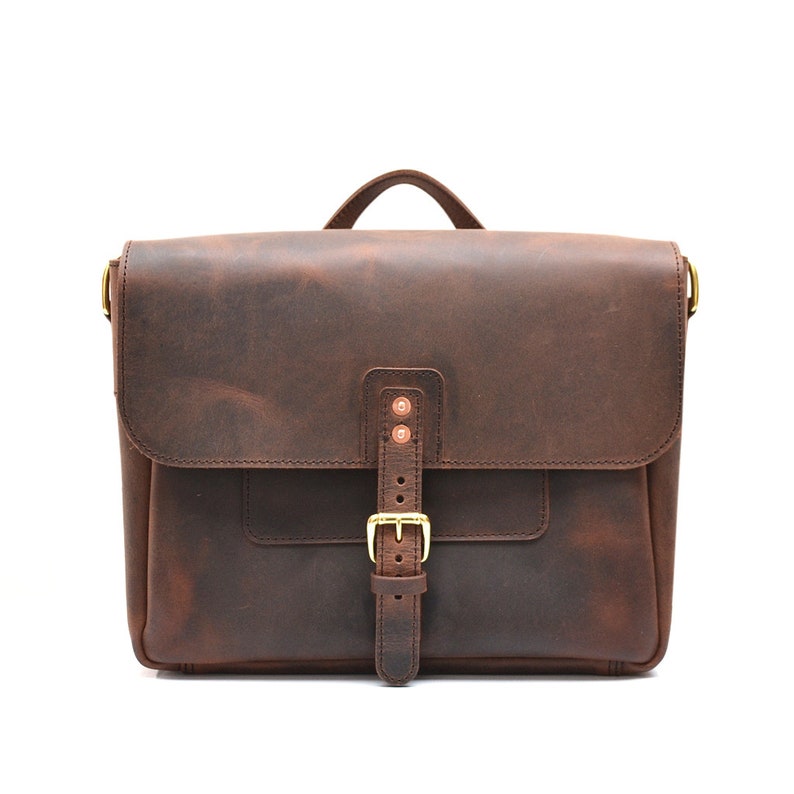 May include: A brown leather satchel with a flap closure and a handle. The bag features a front pocket, a buckle closure, and copper rivets. The leather has a distressed appearance, and the hardware is gold-toned.