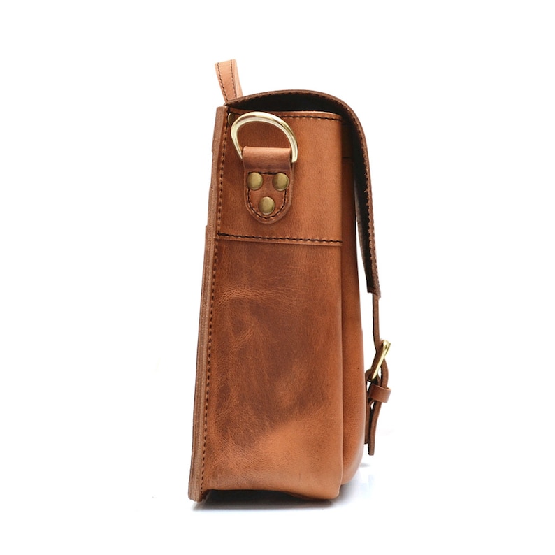 May include: A brown leather messenger bag, side view, with a flap and shoulder strap. It has brass-coloured hardware and visible stitching. The leather has a distressed finish. The bag is designed for carrying items.