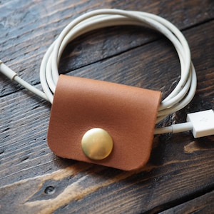 Puede incluir: Un organizador de cables de cuero marrón con un botón de metal dorado. Un cable de carga blanco pasa a través del organizador.