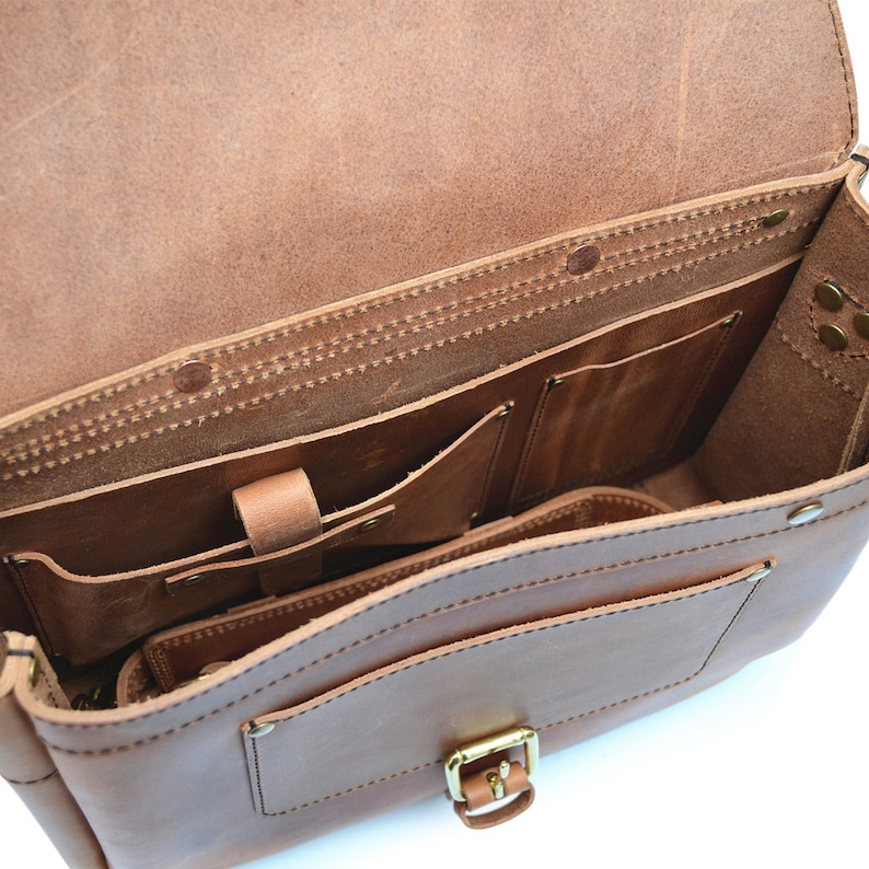 May include: Open brown leather satchel bag with multiple interior pockets. The bag features a flap closure with a gold-coloured buckle. The leather shows visible stitching and a natural grain.