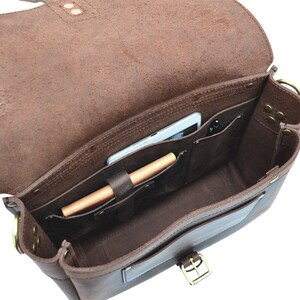 May include: Open brown leather messenger bag with internal pockets. A tablet and pen are visible inside. The bag features a flap closure with a brass buckle and brass hardware. The leather has a distressed finish.
