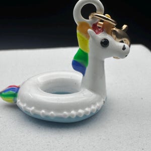 May include: A white and blue unicorn-shaped keyring with a rainbow mane and tail. The keyring has a gold-coloured ring and a gold horn. The unicorn is designed to look like a pool floatie.