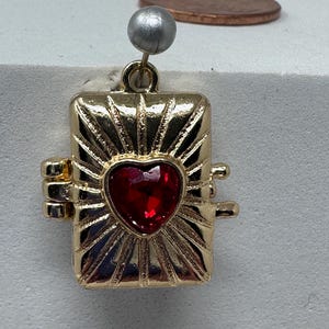 May include: A gold-colored pendant with a red heart-shaped gemstone at its center. The pendant has a rectangular shape with a sunburst design around the heart. A small silver-colored bead is attached to the top.