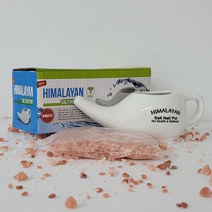 May include: White ceramic Himalayan salt neti pot with a spout and handle. The pot is on a white surface with a small bag of pink Himalayan salt crystals and loose crystals scattered around it. The box for the neti pot is in the background. The box is green and white and has the words "Himalayan Salt Neti Pot" on it.