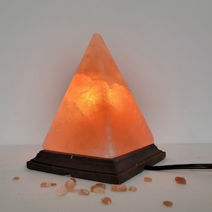 May include: A pyramid-shaped salt lamp with a wooden base, lit and glowing orange. The lamp is surrounded by small pink salt crystals.