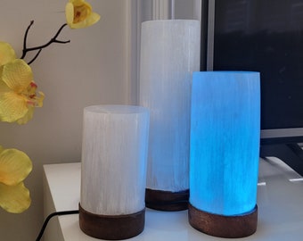 May include: Three selenite lamps, two white and one blue, on a wooden base. The lamps are cylindrical and have a natural, striated texture.