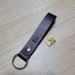 May include: A dark brown leather keychain with a gold key ring and a metal tag with the letter "S" embossed on it. The keychain also has a small gold metal tag with the number "2" embossed on it.