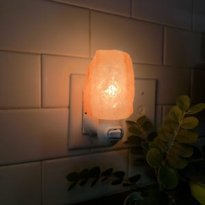 K&ouml;nnte beinhalten: Ein Himalaya-Salz-Nachtlicht, das in eine wei&szlig;e Steckdose gesteckt ist. Die Salzlampe hat eine raue, unregelm&auml;&szlig;ige Form und strahlt ein warmes, orangefarbenes Leuchten aus. Der Hintergrund zeigt wei&szlig;e Fliesen und gr&uuml;nes Laub.