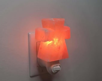 May include: A plug-in night light featuring a carved, translucent orange Himalayan salt crystal in a cross shape. The crystal is mounted on a white plastic base that plugs directly into a wall outlet and includes a small toggle switch.