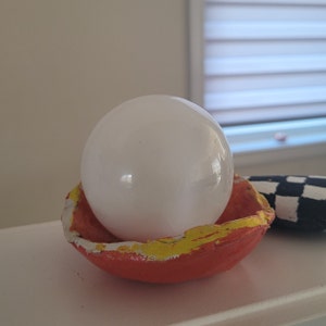 May include: A white sphere sits inside a red and yellow clay bowl. The bowl has a rough, textured surface.